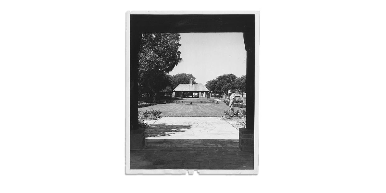 Black and white photo of a man standing in a garden near a small building.