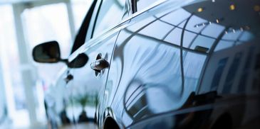 Close-up of a shiny black car door in a showroom reflecting lights and windows.