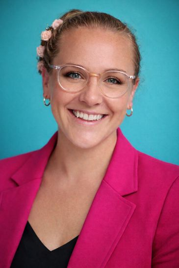 Headshot of Dr Jacinta Thomson, wearing glasses and a pink jacket on a blue background.