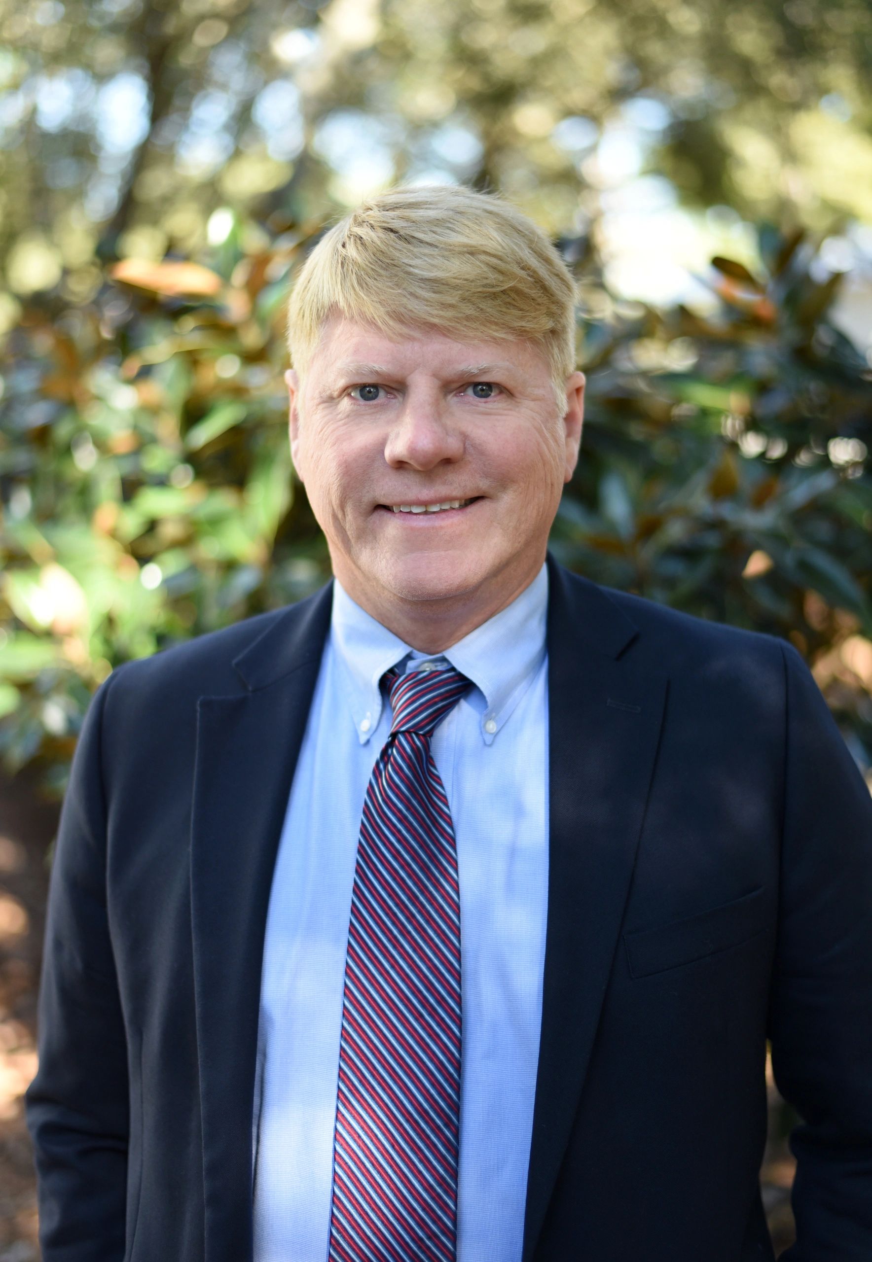 Smiling man in suit and striped tie outdoors with leafy background.