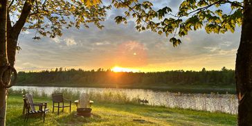 Sunset over a lakeside clearing with chairs and a fire pit.