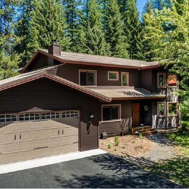 Modern two-story brown house with large garage surrounded by tall green trees.