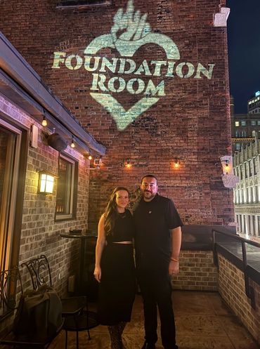 A couple poses under a glowing "Foundation Room" sign on a brick wall at night.