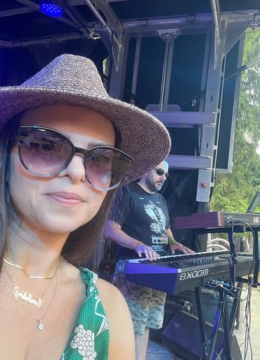 Woman in sunglasses and hat takes a selfie with a man playing keyboard on stage.