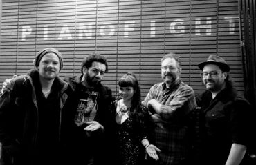 Five people posing in front of a wall with "PIANOFIGHT" sign.