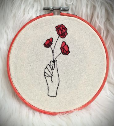 Delicate embroidery of a hand holding three red flowers in a hoop.