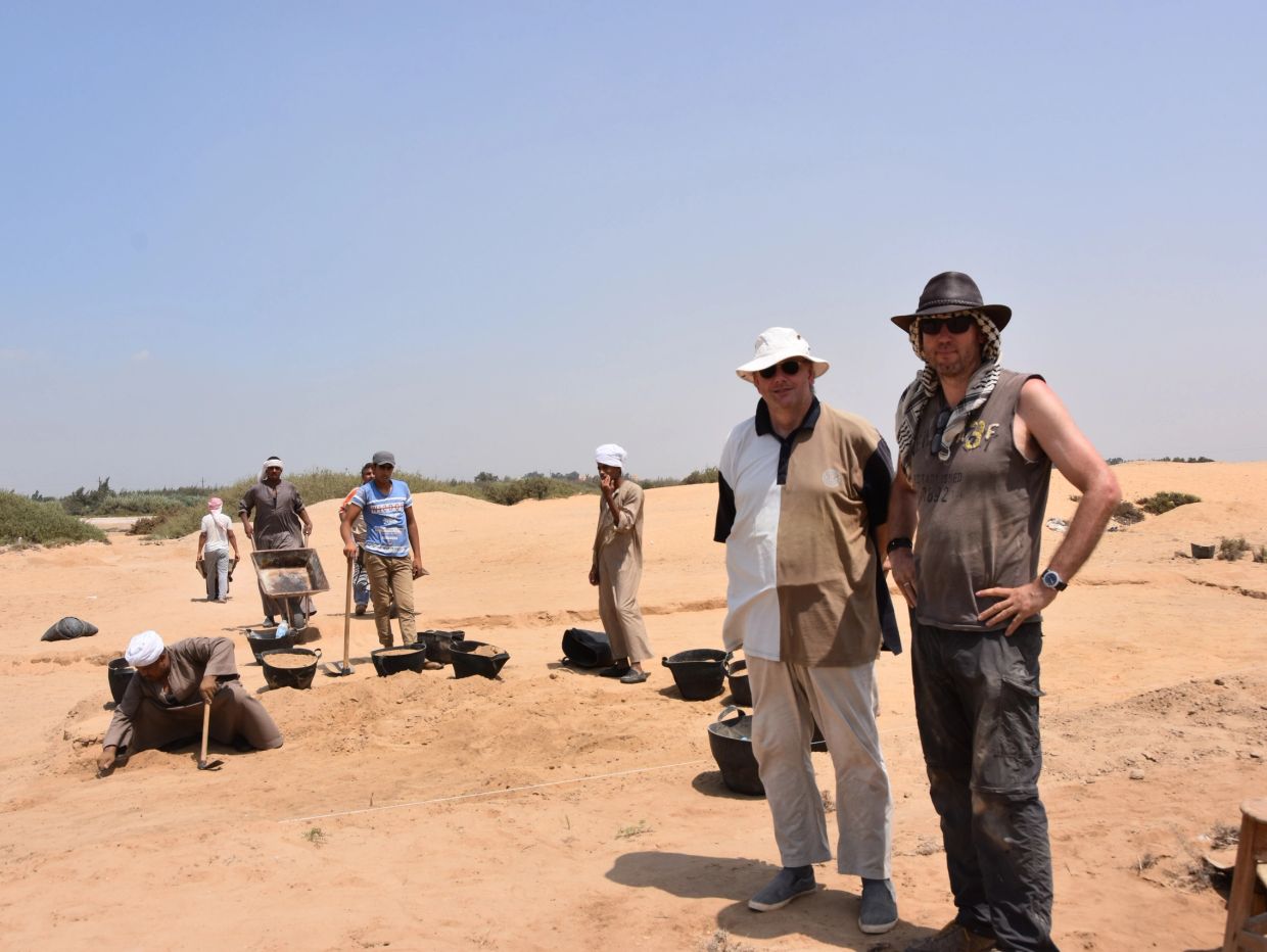 Two men posing at an archaeological site with workers in the background.