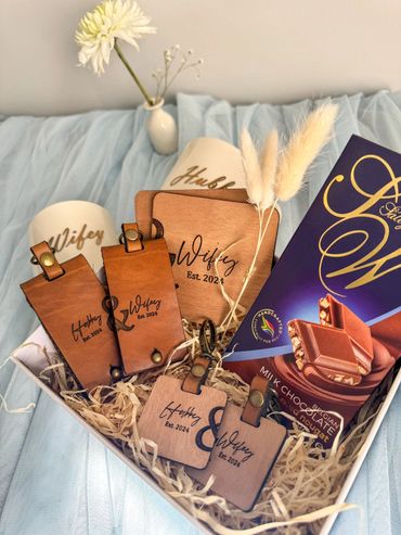 A gift set featuring wooden 'Hubby' and 'Wifey' tags, chocolate, mugs, and decorative flowers.