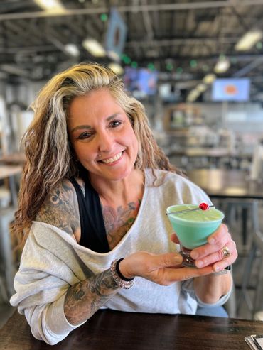 Smiling woman with tattoos holding a blue-green cocktail with a cherry.