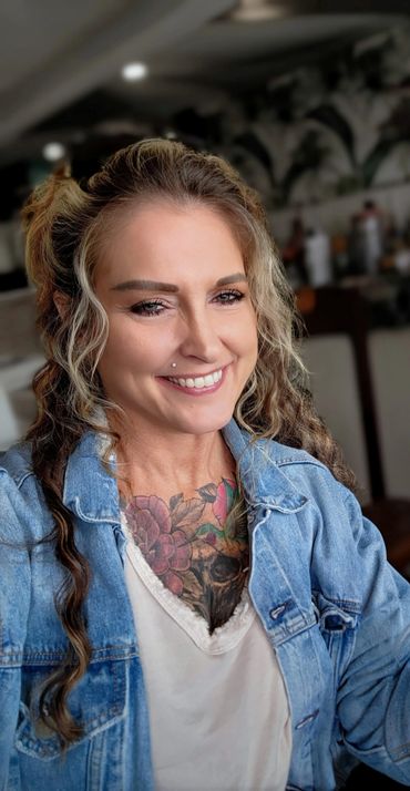 Smiling woman with curly hair, a nose piercing, and colorful chest tattoo in a denim jacket.