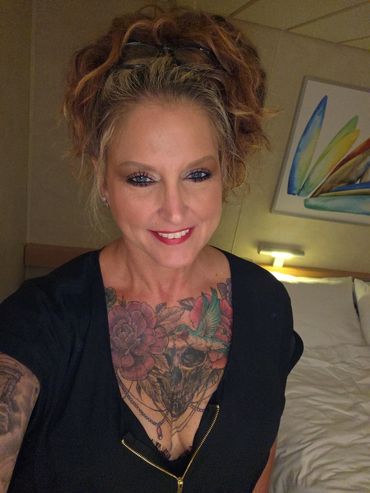 Woman with red hair, a chest tattoo, and a warm smile in a cozy room.