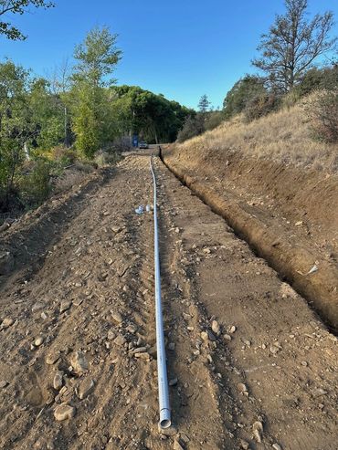 Long PVC pipe laid on a dirt path next to a trench in a rural area.