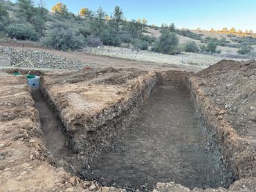 A large rectangular trench is dug in a dirt field near a rocky slope and trees.