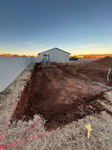 Excavator digging a trench near a white industrial building at sunset.