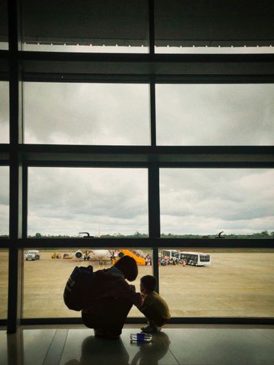 A person and child silhouetted by an airport window with planes outside.
