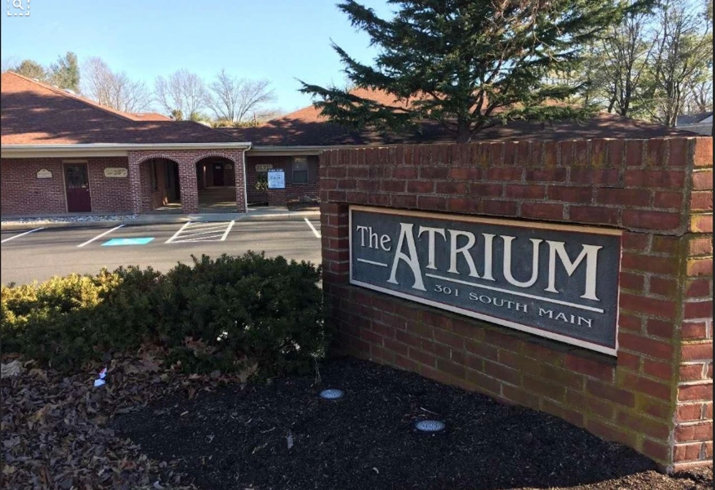 A blue sign with the words "The Atrium 301 South Main" on a brick wall in front of a parking lot.