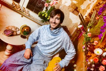 Lächelnder Mann in hellblau gekleidet. Blumen im Hintergrund.