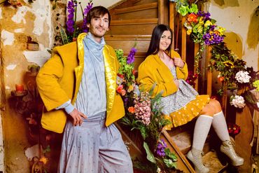 2 Personen vor und auf einer braunen Treppe, beide in Gelb und blau/grau gekleidet. Beide freundlichl