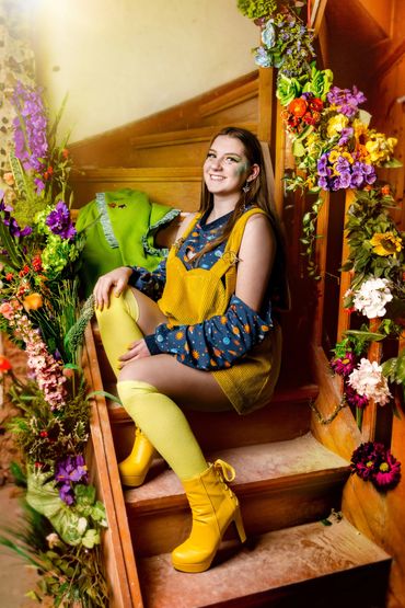 Freundlich lächelnde junge Frau auf brauner Treppe in Gelb und Blau gekleidet.