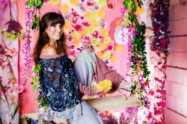 Frau mit braunen langen Haaren auf Schaukel vor einer künstlichen Blumenwand. Sterne voller Blumen.