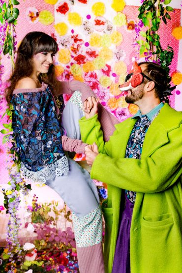 Frau, braune langen Haaren, auf Schaukel vor einer Blumenwand, ihr zugewand ein Mann mit Fuchsmaske.