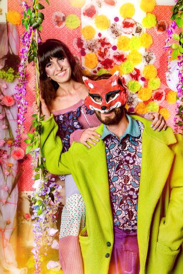 2 Personen vor einer Blumenwand. Eine Frau sitzend auf einer Schaukel ein Mann davor mit Fuchsmaske.