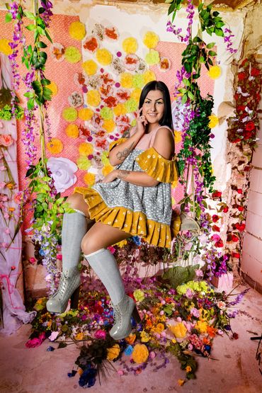 schwarzhaarige junge Frau mit kurzem Kleid auf einer Schaukel vor Blumenwand.