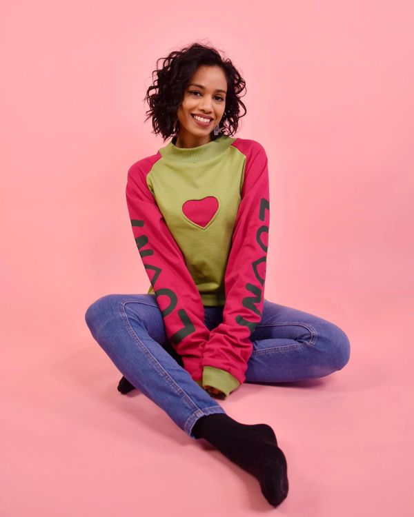 Frau mit dunklen Haaren und breitem lächeln sitzt vor rosa Hintergrund, bekleidet mit Jeans und Pulli