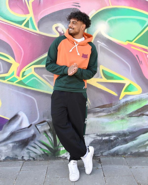 Cooler Typ vor Grafitti mit Orange/grünem Pulli, schwarz/grüner Jogginghose und schwarzen Locken.