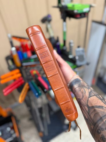 A hand holding a brown leather-bound Holy Bible with a tattooed arm.