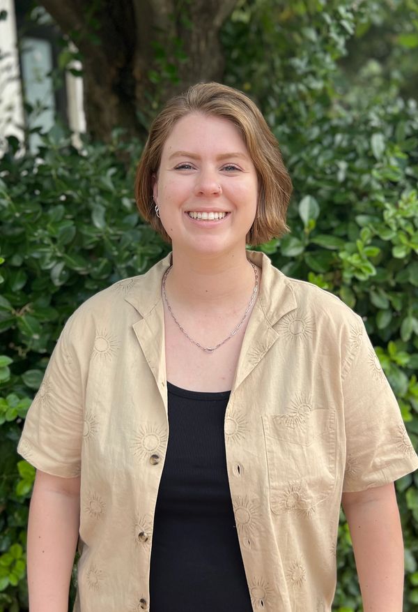 Smiling person in a beige shirt stands outdoors with greenery in the background.