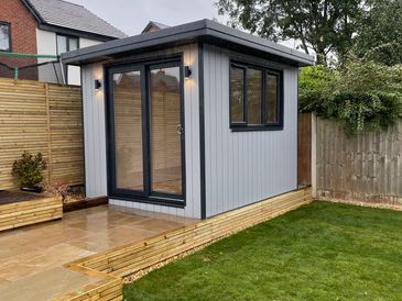 Modern timber-clad garden building with grey accents on paved patio and lawn backdrop.