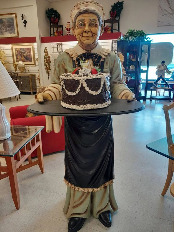 Statue of an elderly woman holding a tray with a chocolate cake in a vintage-style room.