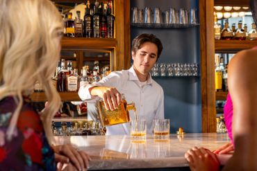 Bartender pouring whiskey into glasses at a bar with two women.