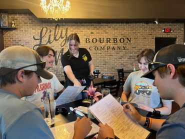 Four young men sit at a table with menus while a waitress smiles and points at one.