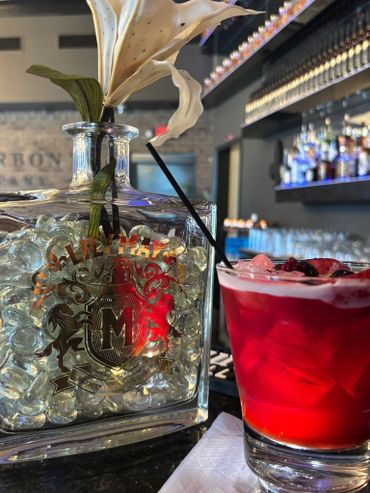 A vibrant red cocktail beside a decorative glass bottle with a flower inside, set on a bar counter.
