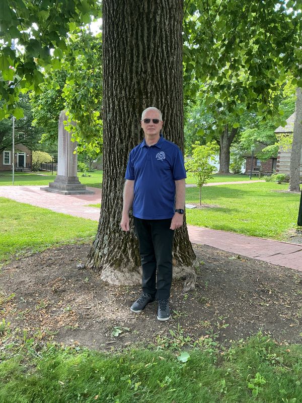 Man wearing sunglasses and blue polo shirt standing by a large tree in a park.