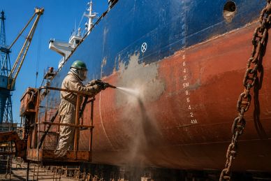 Worker power washing a ship's hull in a dry dock.