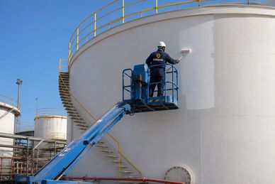 Worker painting large industrial storage tank white using a lift platform.