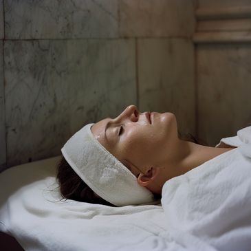 A woman relaxing with eyes closed, wearing a white headband and robe in a spa setting after a relaxation massage.