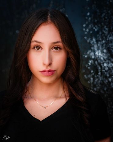 Portrait of a young woman with brown eyes and long hair, wearing a black top and a gold necklace.
