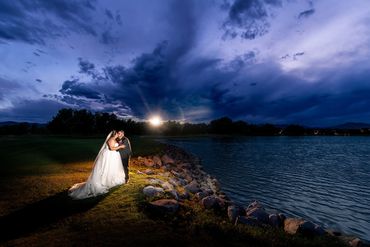 A newlywed couple shares a kiss by a lakeside at dusk with dramatic clouds overhead.