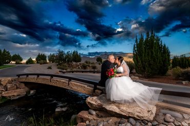 Bride and groom kiss on a stone bridge under a dramatic sky.