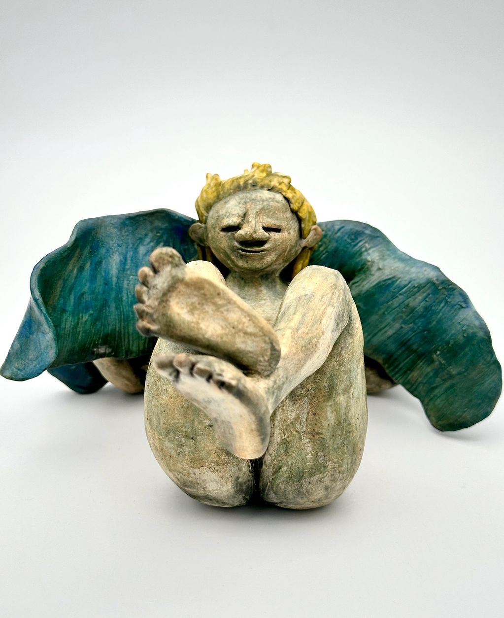 A whimsical winged figurine with yellow hair sitting with legs extended.