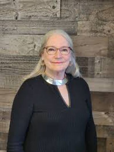 Smiling elderly woman with glasses and silver necklace against a wooden wall.