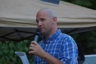 Man in blue plaid shirt speaking into a microphone outdoors.