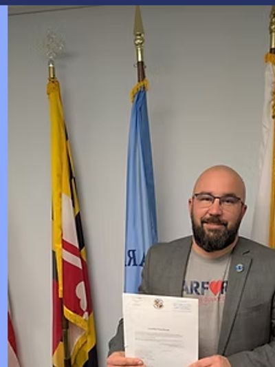 Man in blazer holding a document, standing between flags indoors.