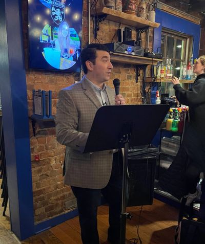 Man in plaid blazer speaking into a microphone at a bar or restaurant.