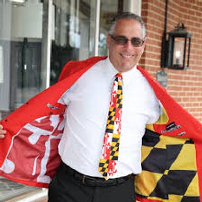 Man showing the inside of his red jacket with a Maryland flag lining and wearing a Maryland-themed tie.