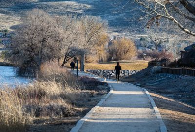 Two people walk along a paved path by a river in a scenic, quiet area.